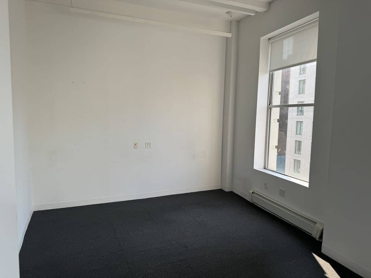 A small, empty office with white walls, a large window, and dark gray carpet.