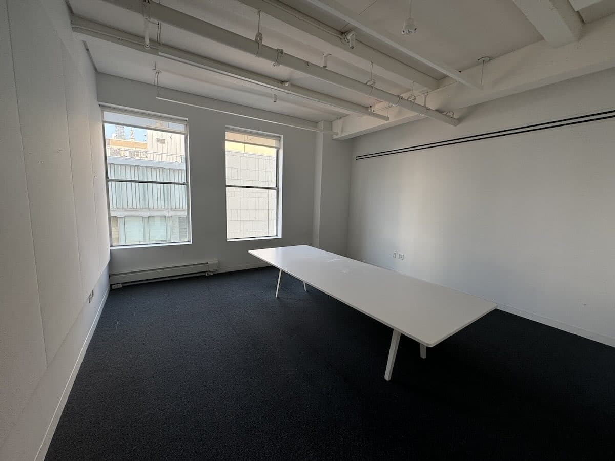 A minimalist office with white walls, black carpet, and large table.