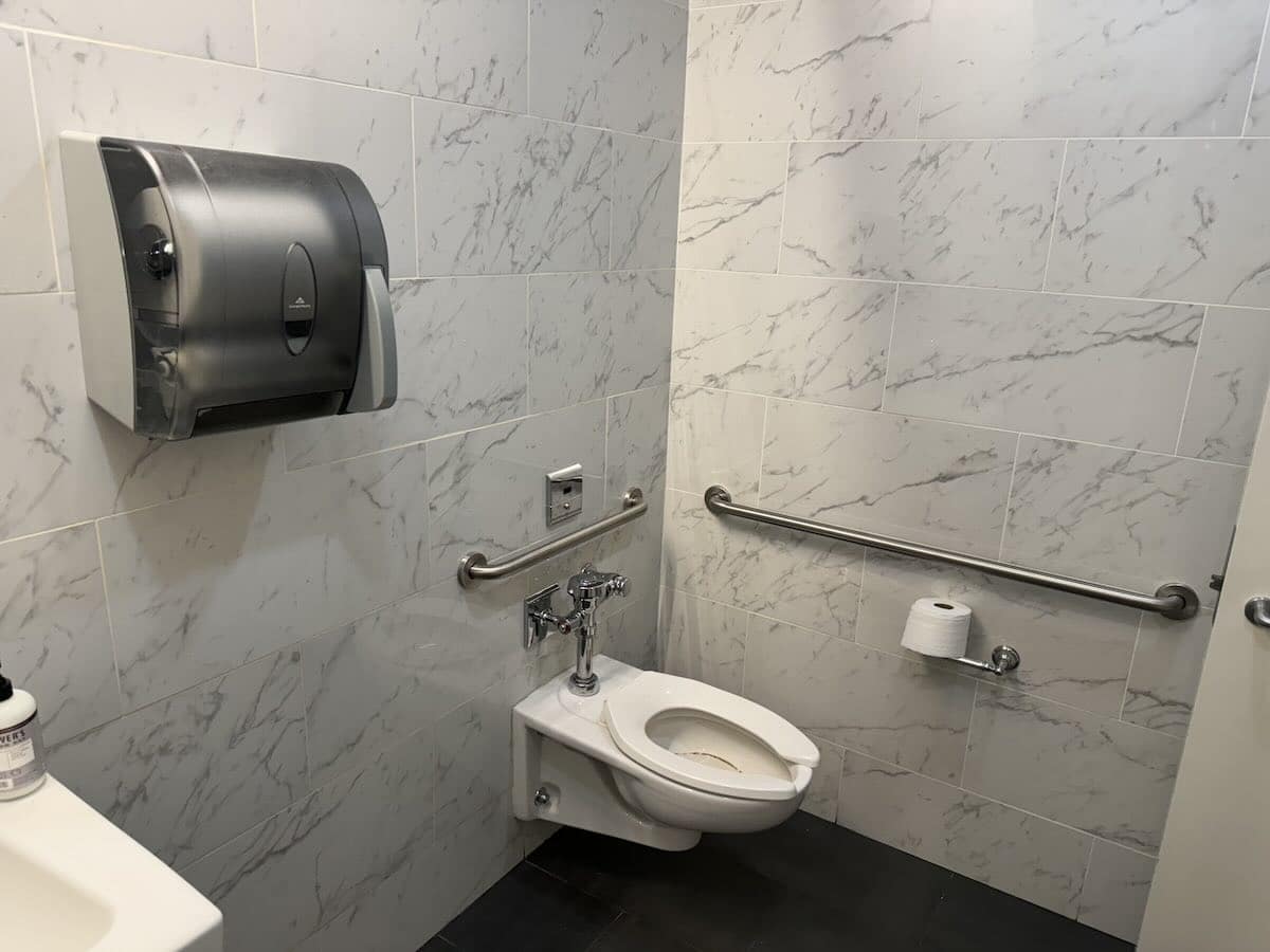 Public restroom, light gray tiles, wall-mounted toilet.