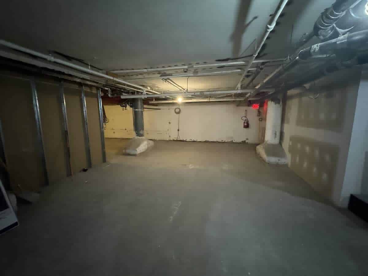 ALT tag: Unfinished basement with exposed pipes, bare concrete floor, and minimal light.