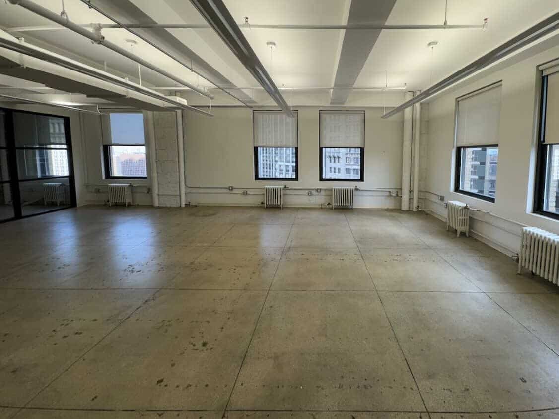 Empty office with large windows, exposed pipes, and polished flooring.