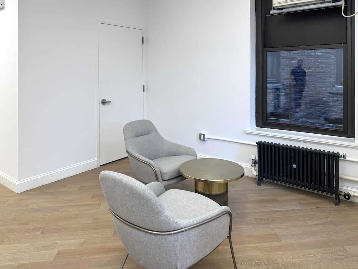 A room with armchairs, gold table, wood floor, door, and a large window