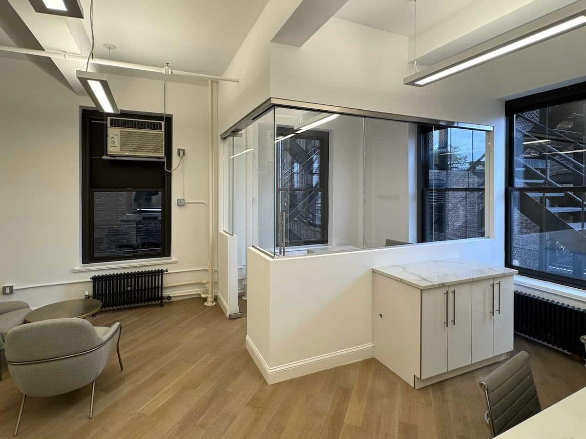 Office with glass partitioned room, gray chairs, AC unit, shelving, large windows, and hardwood floor.