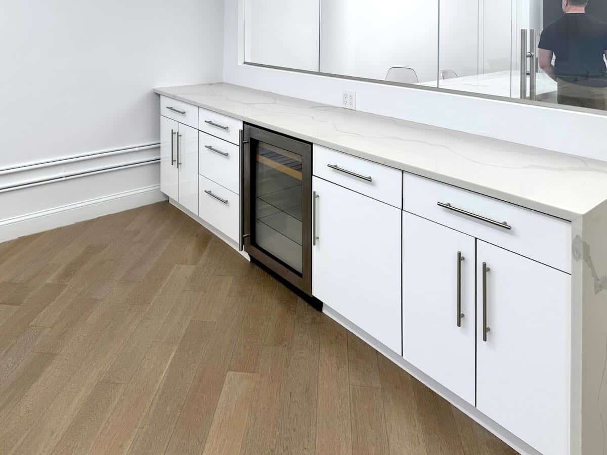 A mini pantry with white cabinets, light wood flooring, and a wine cooler.