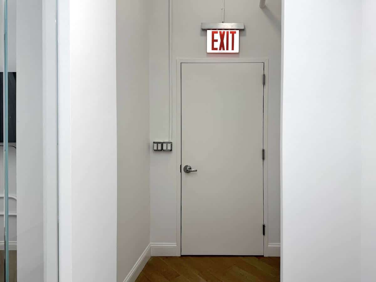 White hallway with wooden floors leading to a closed white door with an exit sign