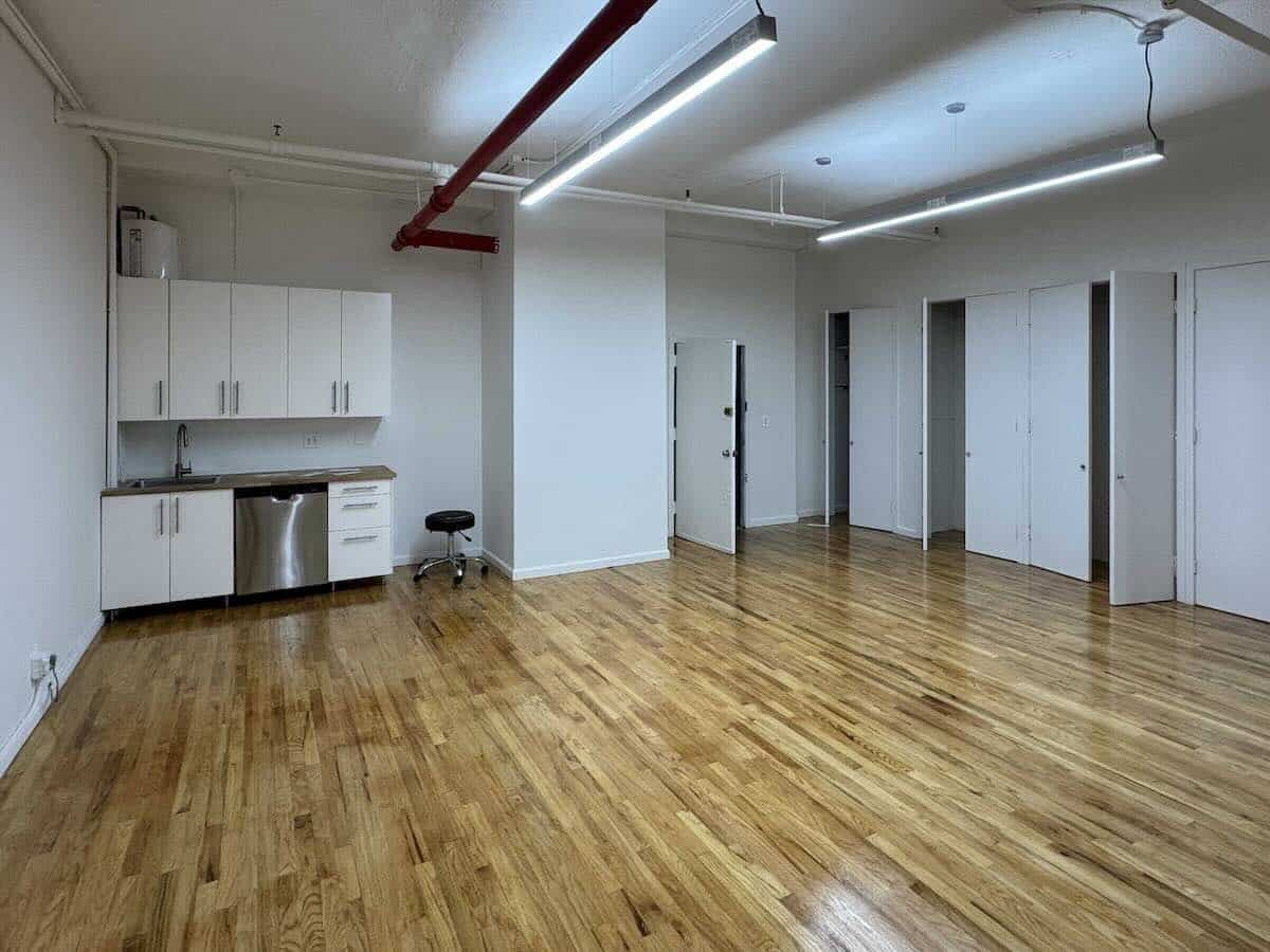 Spacious empty room with polished floors, white walls, and kitchen area.