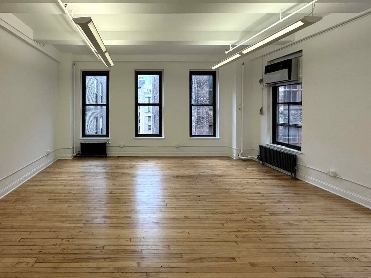 A spacious, empty room with wood floors, white walls, and large windows.