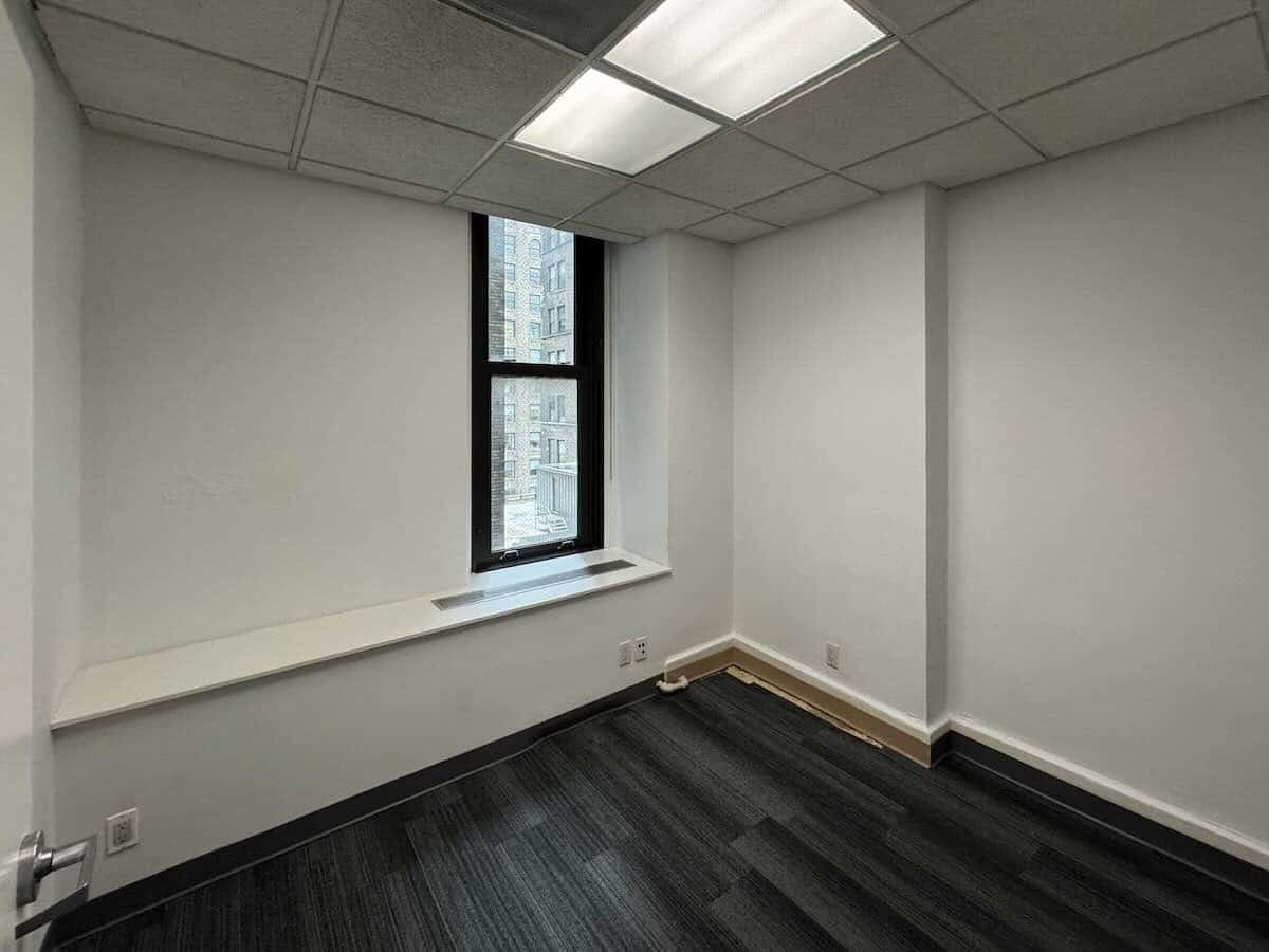 Empty office with window, white walls, black carpet, and wall shelf.