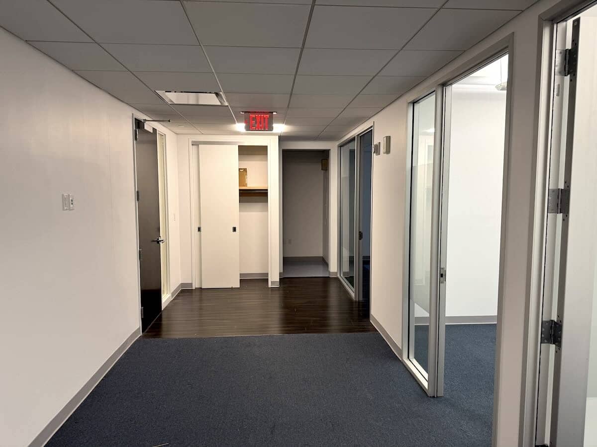 Office hallway with white walls, glass partitioned rooms, wood and carpet flooring.