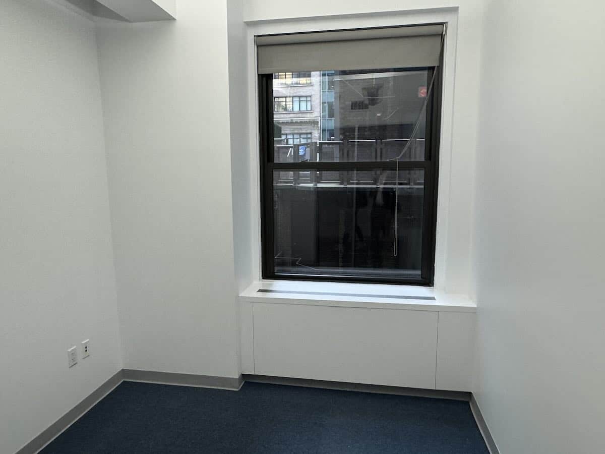 Empty office with white walls, dark blue carpet, a large window with a grey blind.