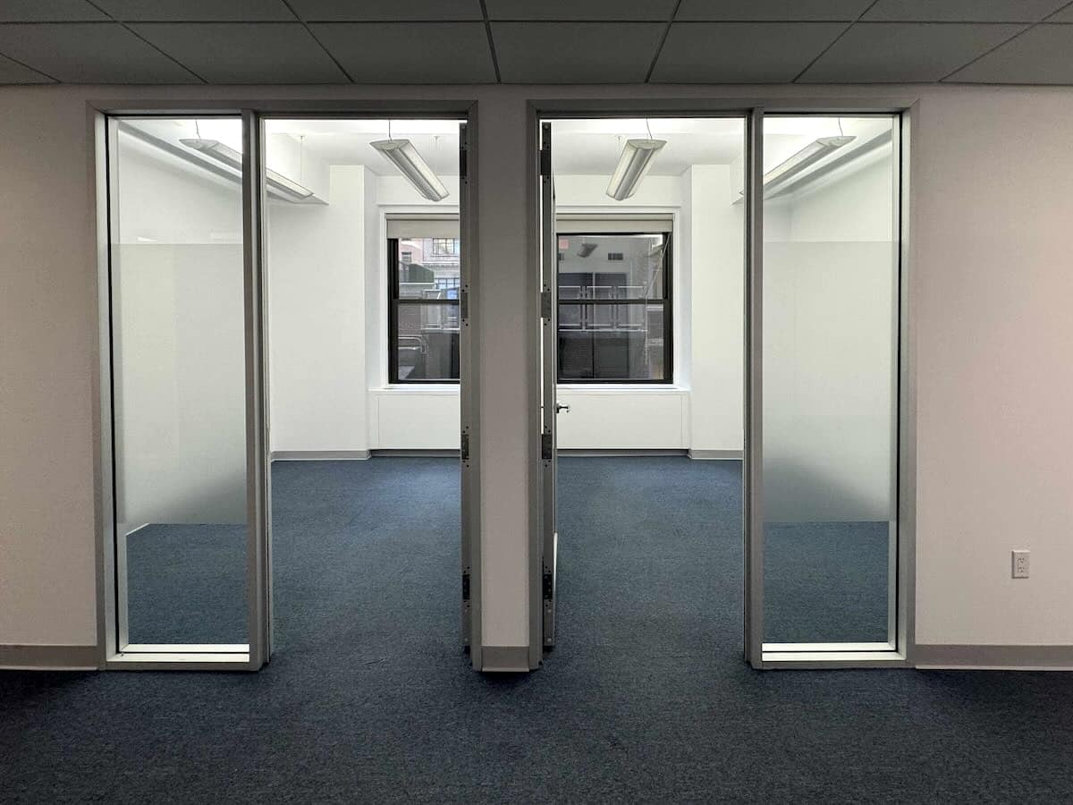 Modern empty office with glass enclosed rooms, gray carpet, and large windows.