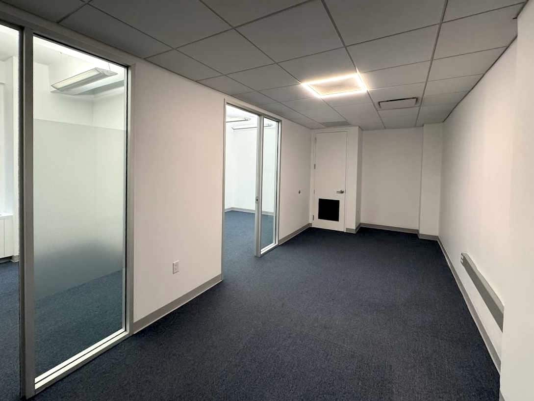 A modern, empty office with white walls, dark grey carpet, and glass partitions.