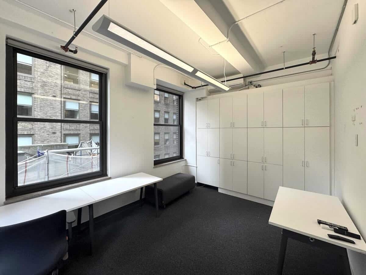 Small office with large windows, white walls, desks, and chairs.