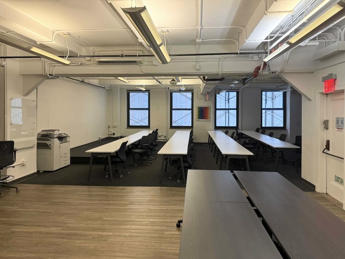 Modern office with tables, black chairs, pipes, and large windows.