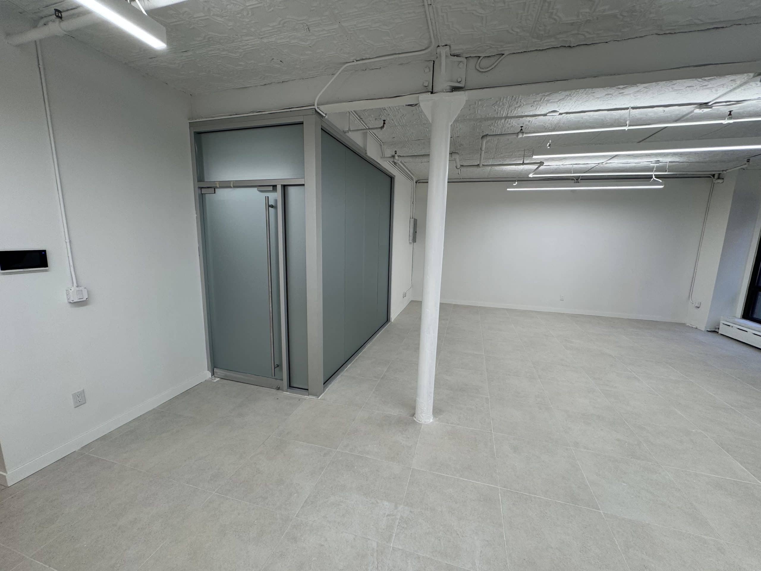 Empty room with tiled floor, white walls, glass-panel area, ceiling pipes.