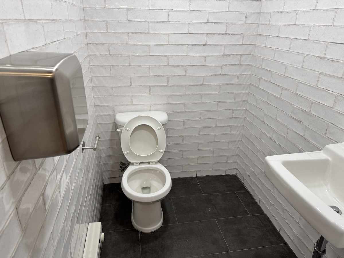 White brick bathroom with dark tiles, toilet, sink, and dispenser.